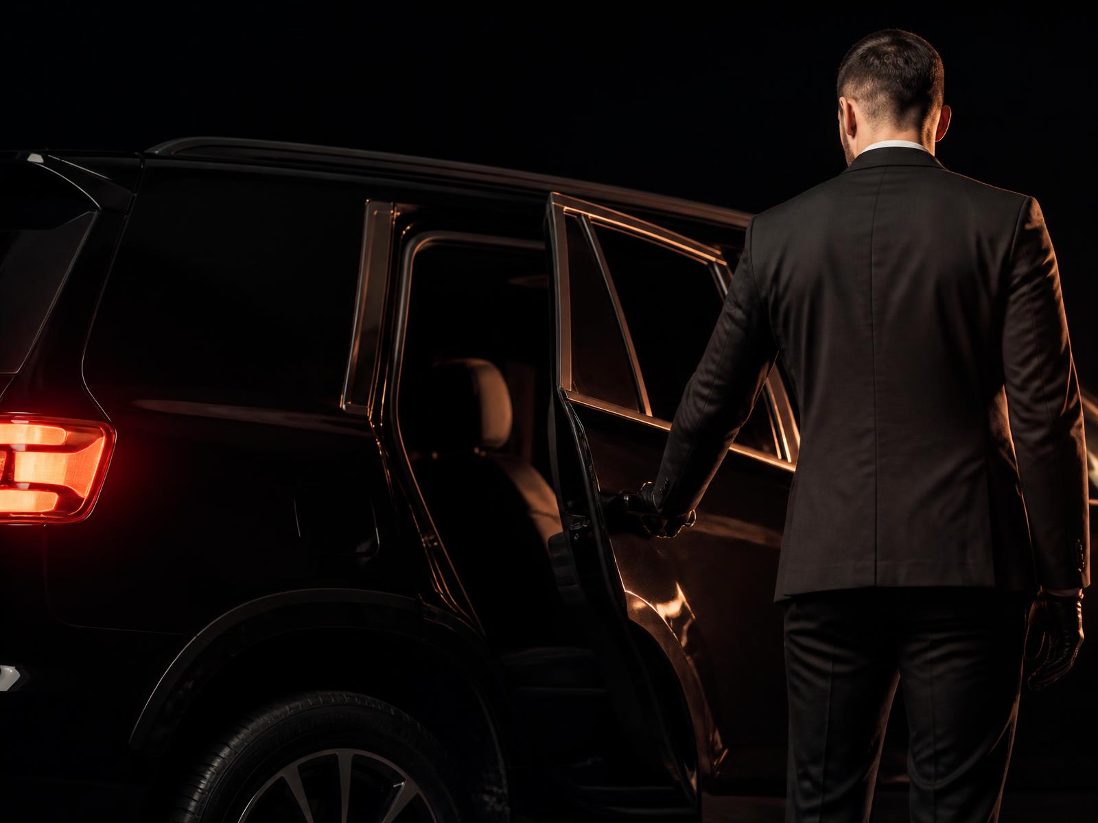 Professional chauffeur opening the rear door of a black luxury SUV at night