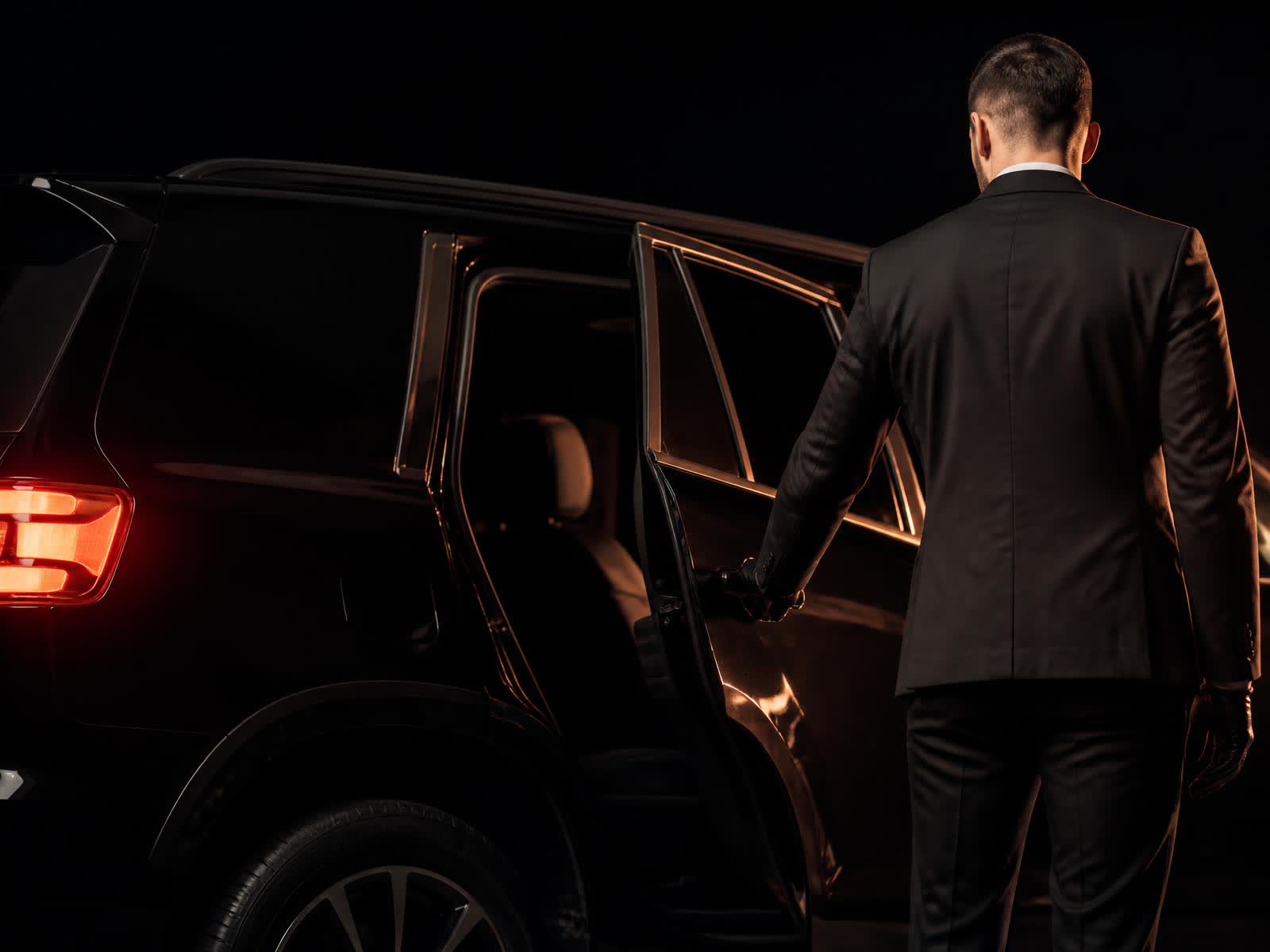 Professional chauffeur opening the rear door of a black luxury SUV at night